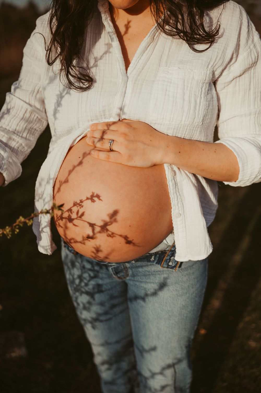 Pregnant woman in white shirt and jeans exposing baby bump with floral shadow in warm golden light.