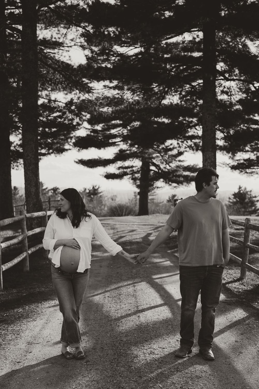 Black and white photo of a pregnant couple holding hands on a tree-lined path with a wooden fence.