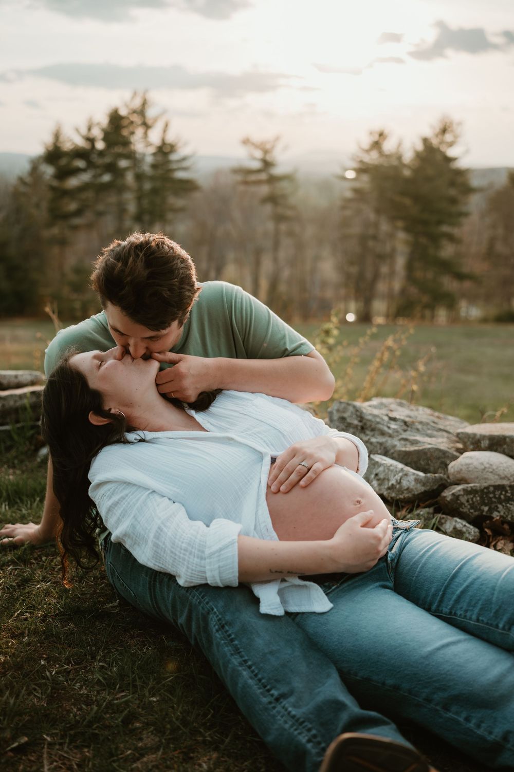 Couple shares a tender kiss outdoors, pregnant woman lying on grass against stone wall at golden hour.