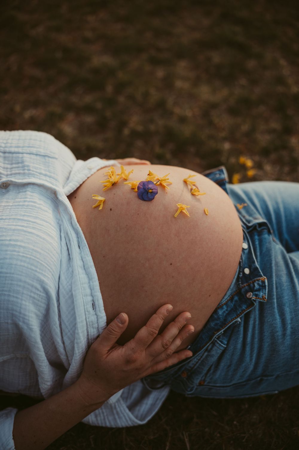 Pregnant woman lying on grass with yellow and purple flowers resting on her bare baby bump, wearing jeans and white shirt.
