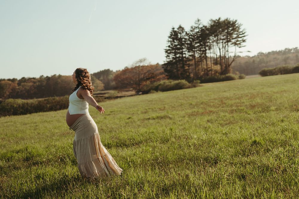 Pregnant woman in white top and beige skirt standing in a sunlit green field with trees in the background.
