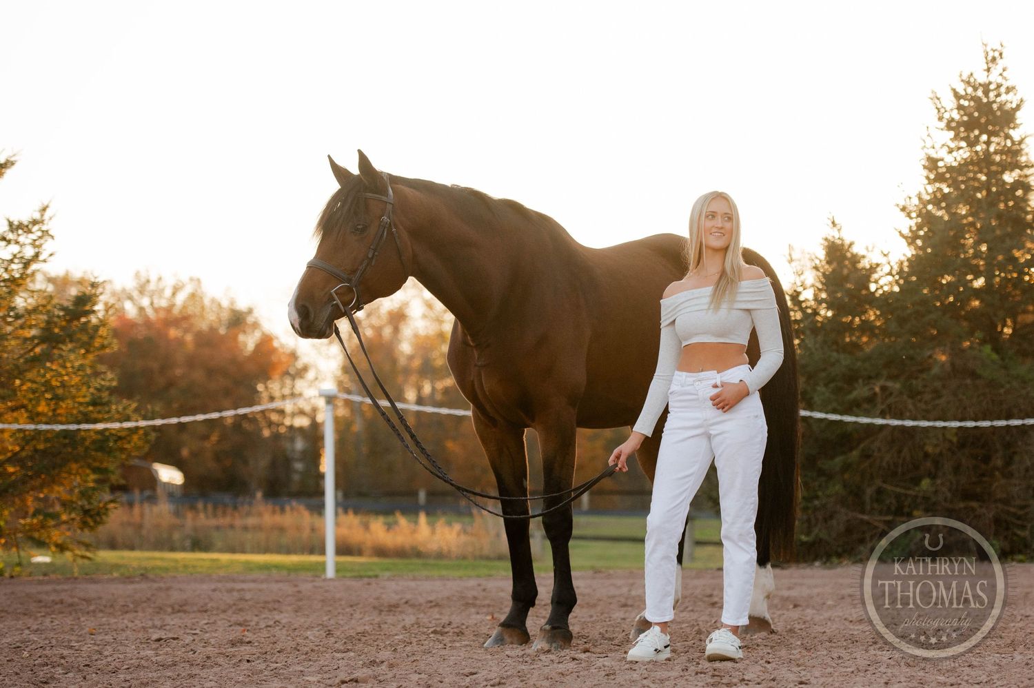 A person and dark horse stand together in a fenced arena at sunset.