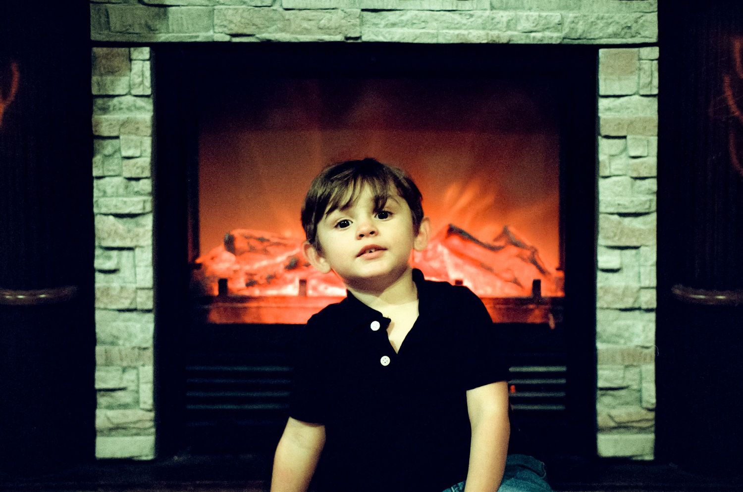 Child in black shirt sitting in front of a glowing electric fireplace with stone surround.