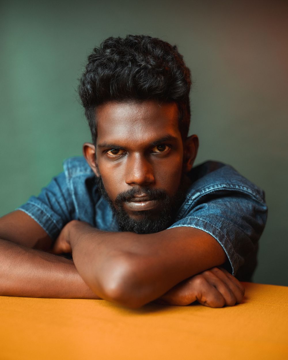 Portrait photo against green background showing person in denim jacket resting arms on orange surface.