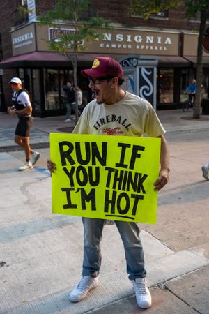 Someone holds a bright yellow sign reading 'RUN IF YOU THINK I'M HOT' on a city street near storefronts.