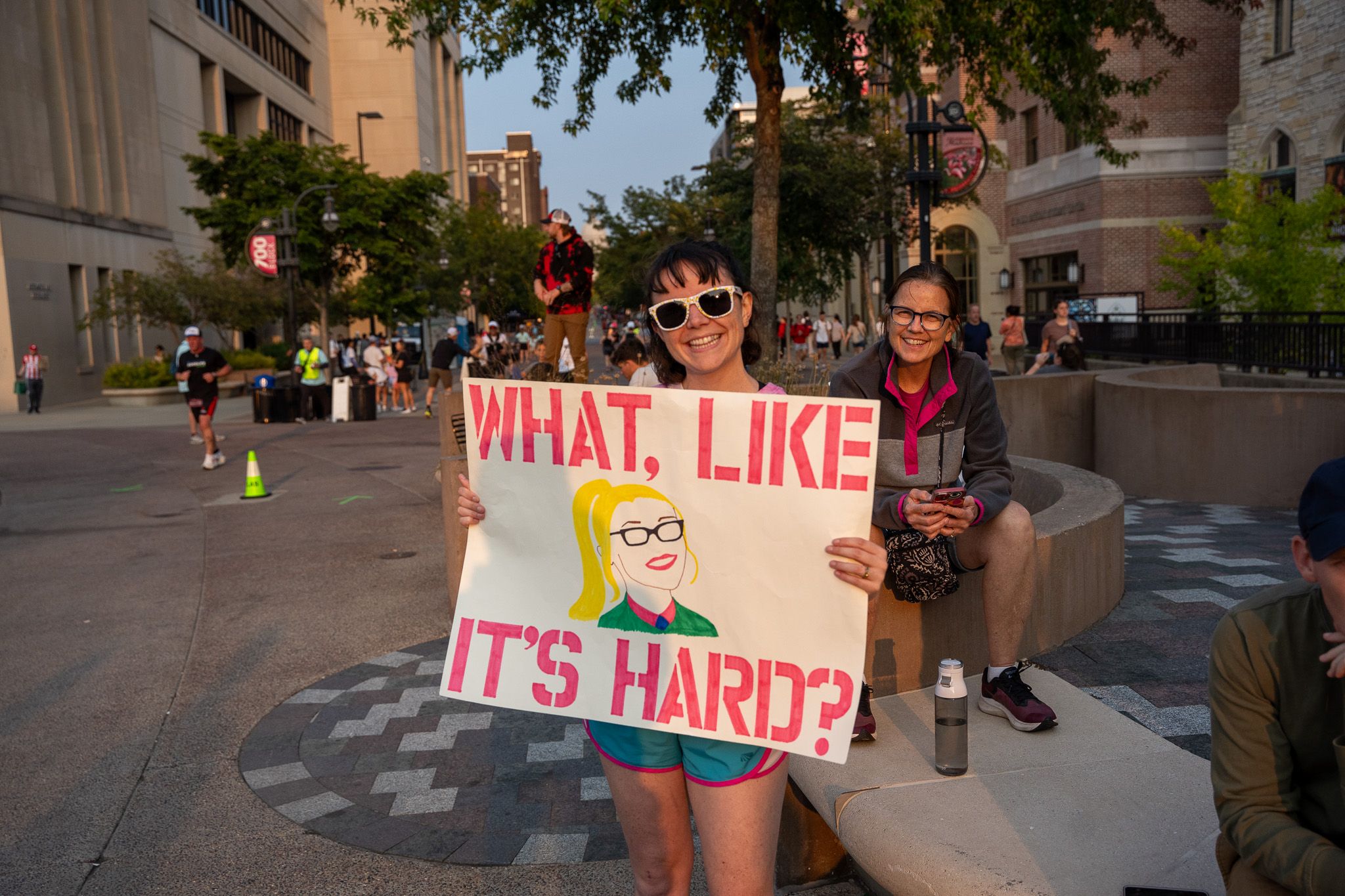 A group of people pose on a city street with a sign displaying 'WHAT LIKE IT'S HARD?' in pink lettering.