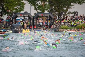 Triathletes swim in open water during a race with colorful swim caps and large crowds watching from shore.