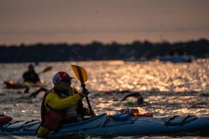 A kayaker paddling at sunset on a glittering water surface with safety gear and equipment visible.