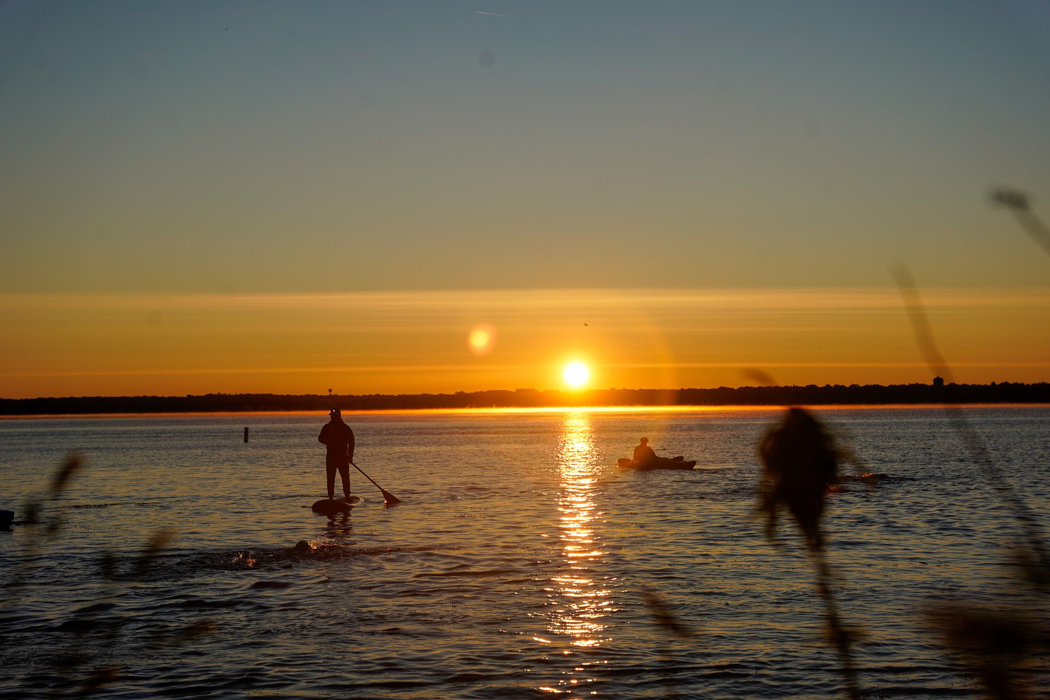 A paddle boarder silhouetted against a vibrant orange sunset over calm waters with golden sun reflection.