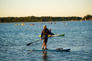 A paddleboarder stands on calm blue water at sunset with kayakers visible in the background.