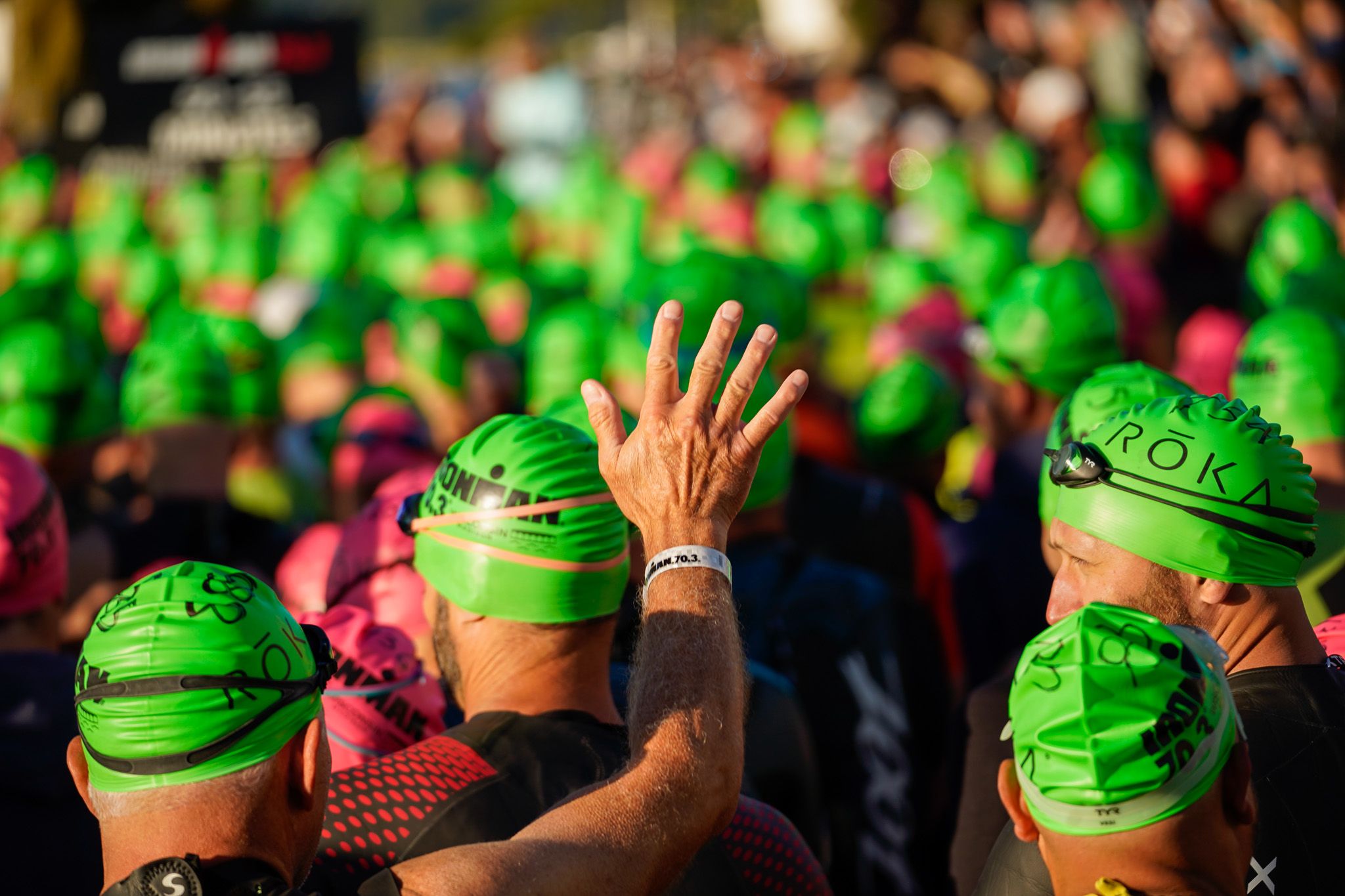 Crowd of athletes wearing bright green swim caps with raised hand at a triathlon event.