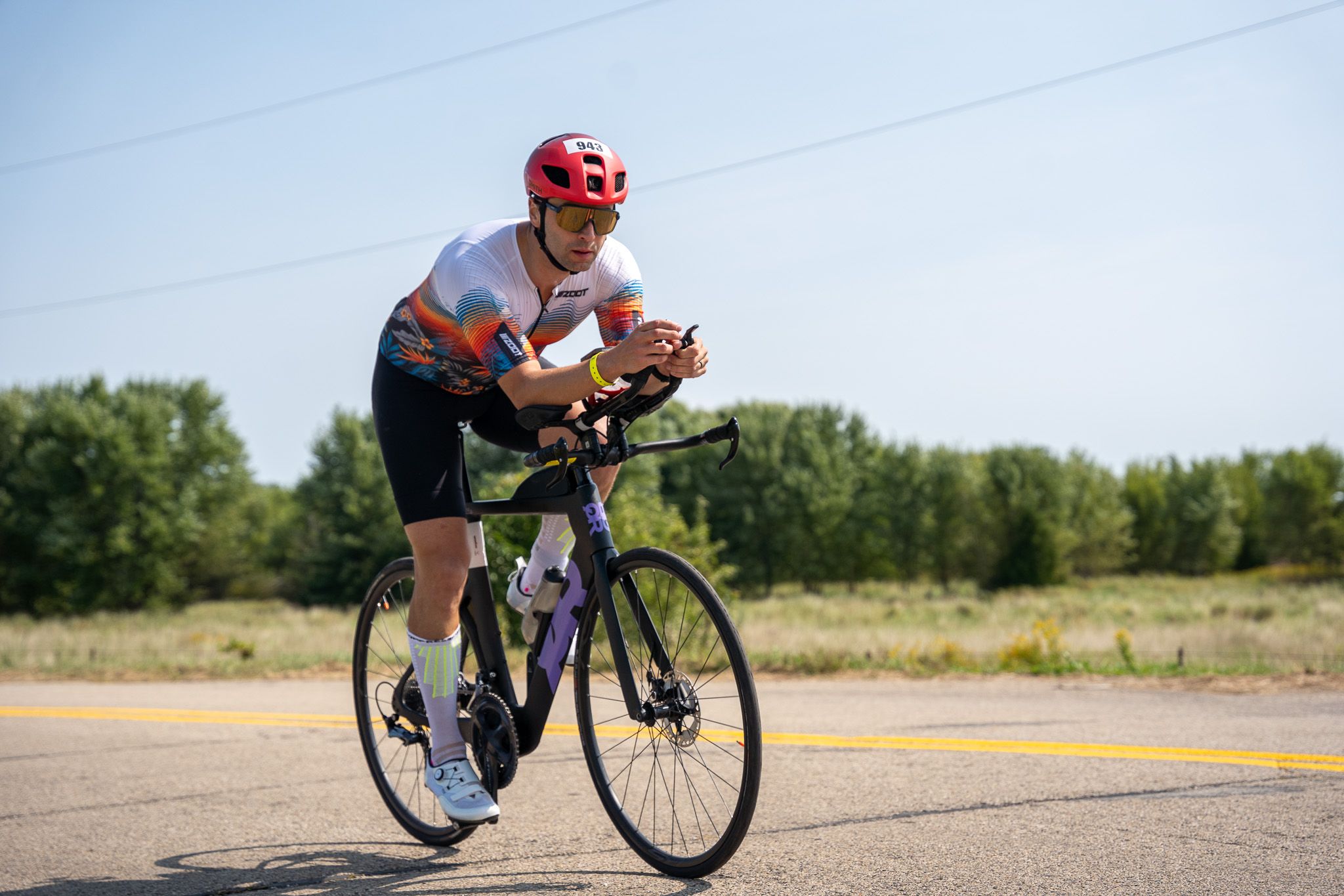 A cyclist in red helmet and athletic gear rides a time trial bike on a rural road on a sunny day.