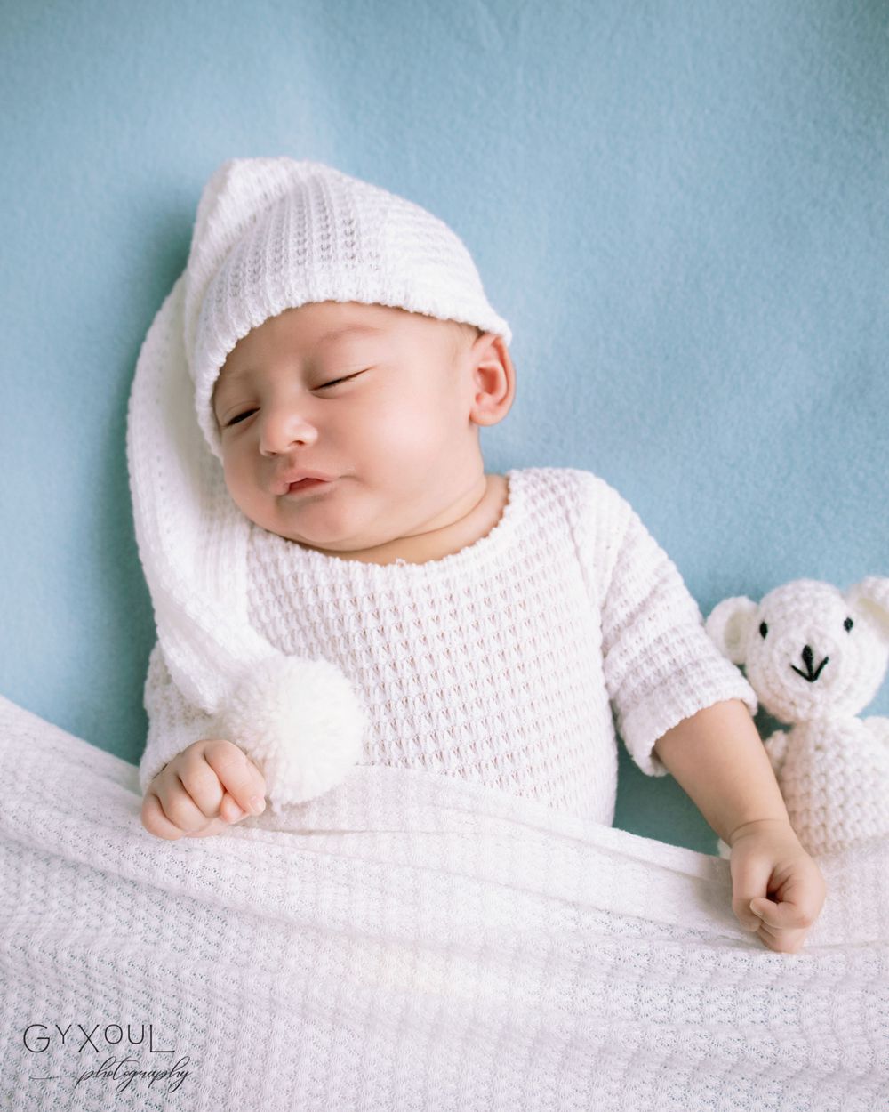 Newborn photo in white knit outfit sleeping beside small plush toy against blue background.