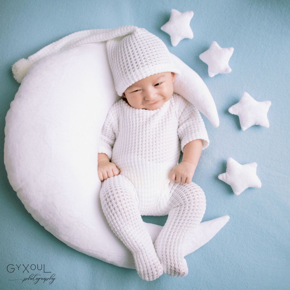 Newborn photo  in white knitted outfit resting on crescent moon prop with decorative stars.