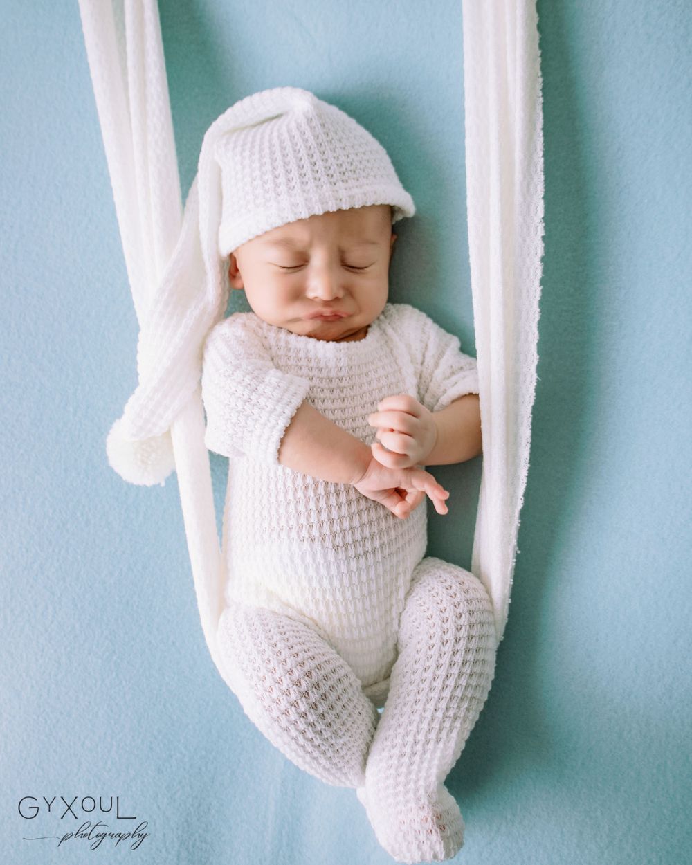 Newborn photo  in white knitted outfit sleeping peacefully in hanging fabric swing.