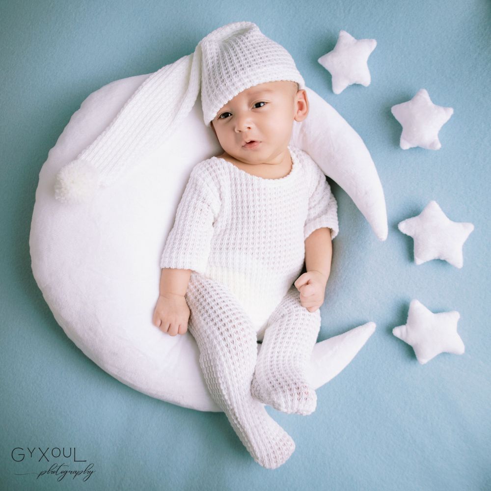 Newborn photo  in white knit clothing posed on moon-shaped cushion with star decorations.