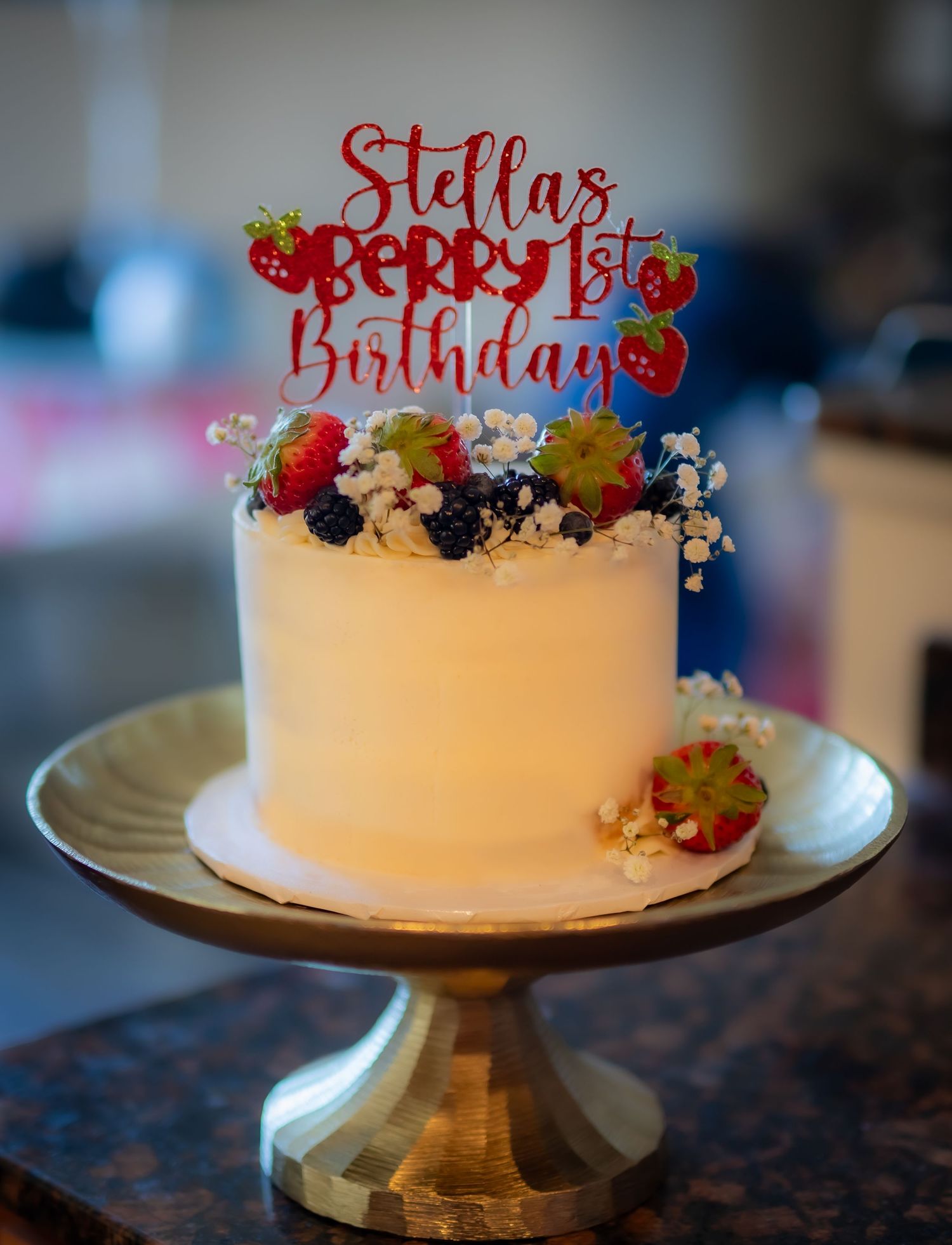 Birthday cake with red berry topper and fresh fruit decorated in white frosting on a metal cake stand.