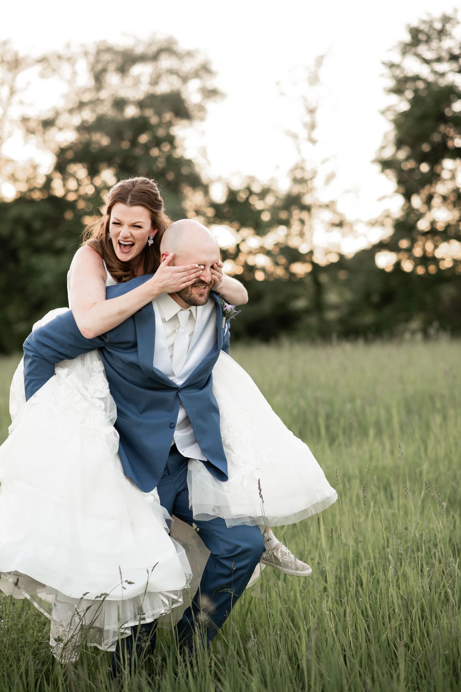 A playful wedding photo in a grassy field at sunset showing joyful couple laughing and embracing outdoors.