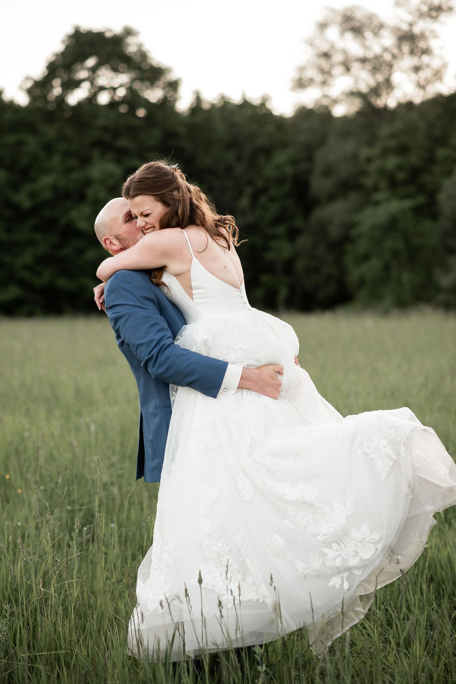 A romantic wedding couple embraces in a green meadow at dusk with the bride's white dress flowing in the breeze.