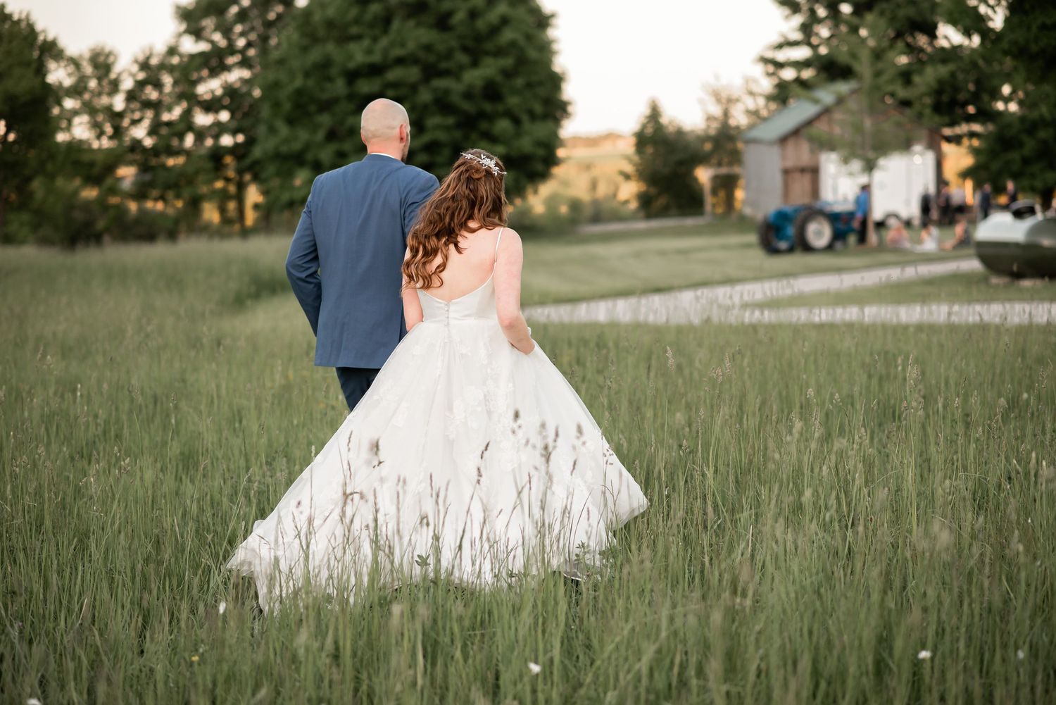 A couple in wedding attire walks through a lush green meadow at sunset with a farm in the background.