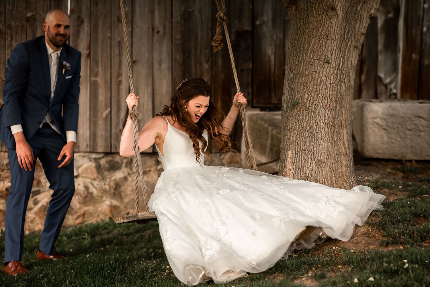 A bride in a flowing white wedding dress swings playfully on a rope swing outside a rustic wooden building.