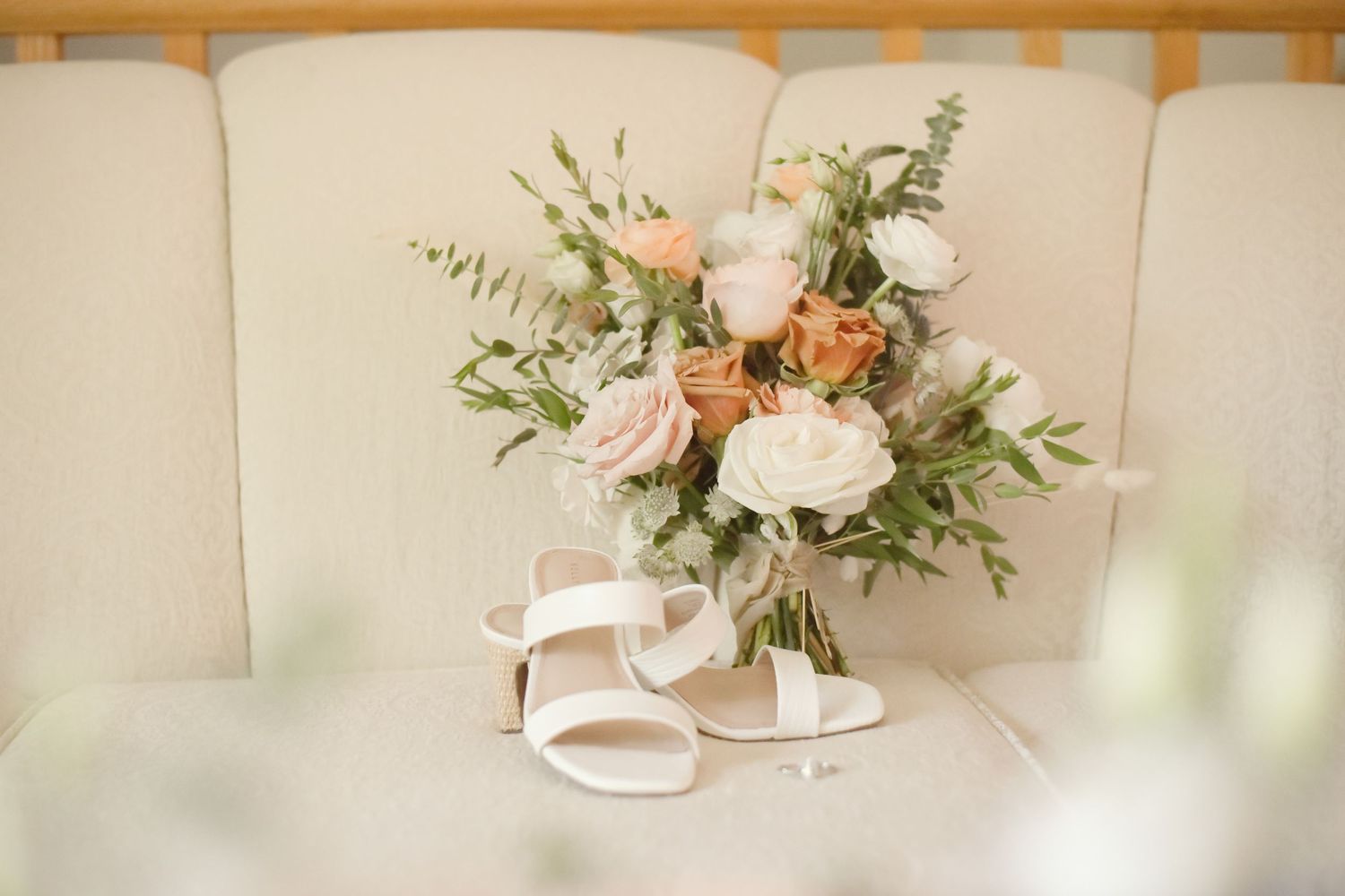 Elegant bridal bouquet of pale pink and peach roses with greenery rests on white tufted cushion beside white sandals.
