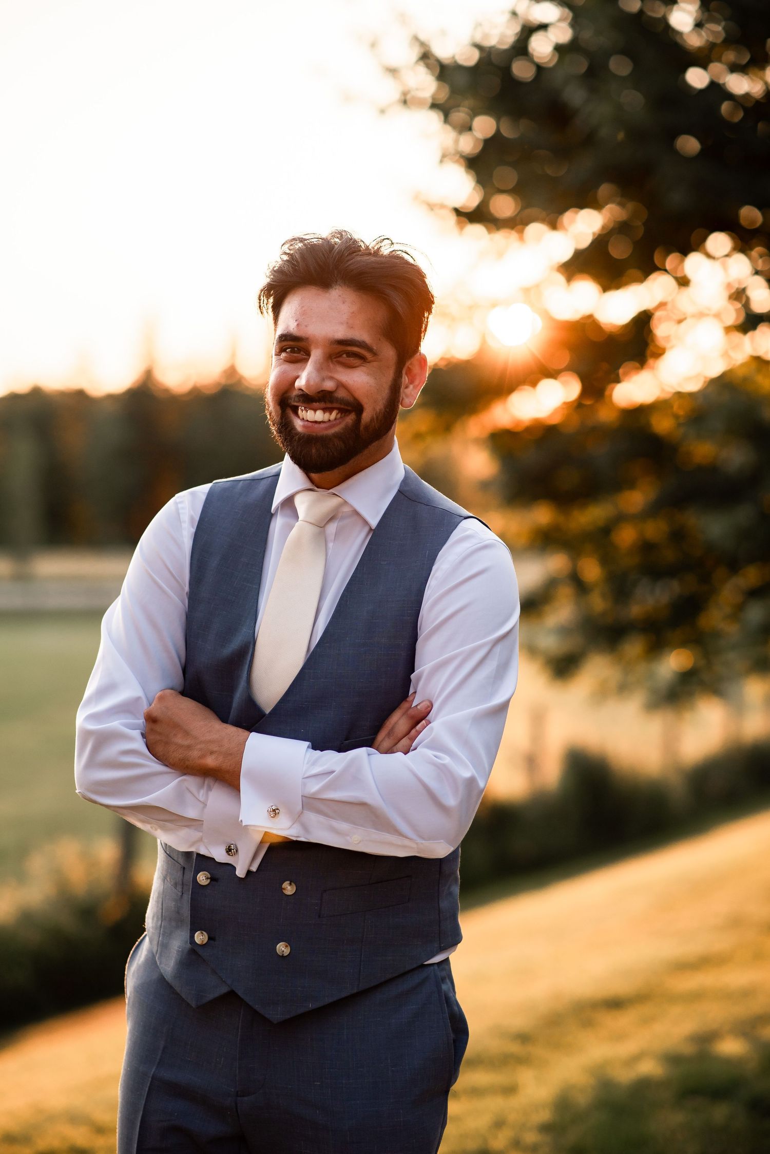 A person wearing a blue vest and white dress shirt smiles warmly against a golden sunset backdrop in an outdoor setting.