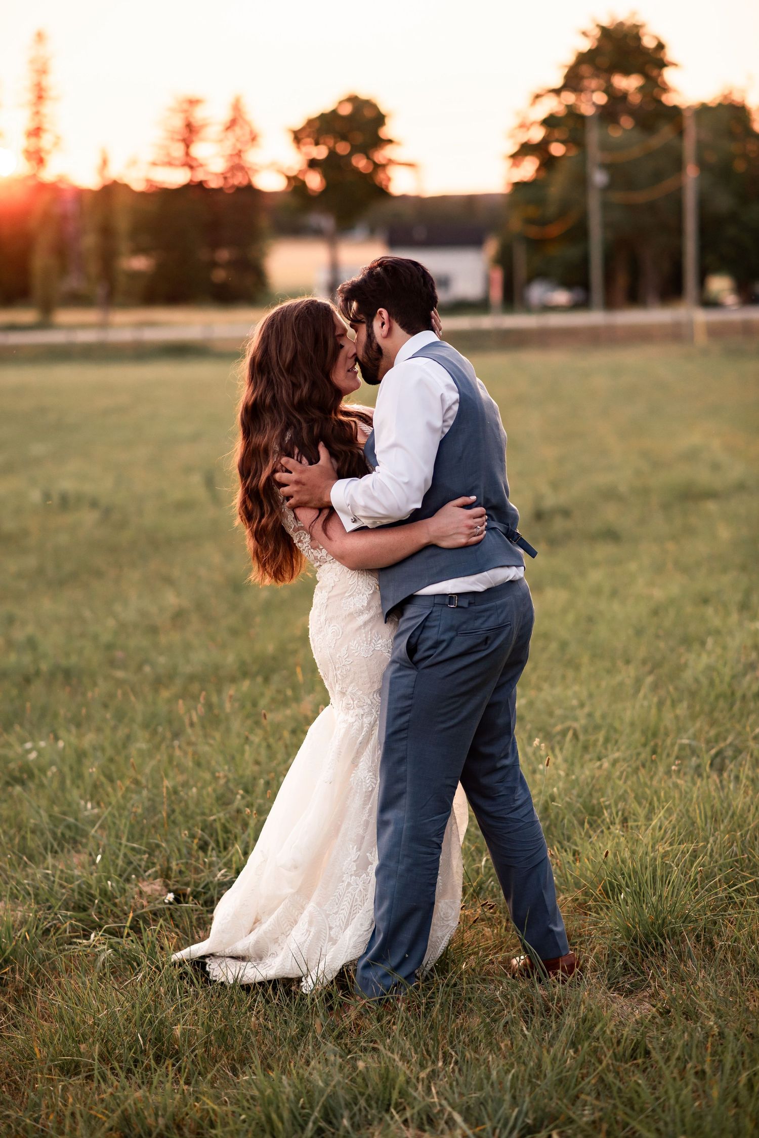 A couple shares a romantic embrace in a grassy field during golden hour sunset, the bride wearing a flowing lace wedding gown.
