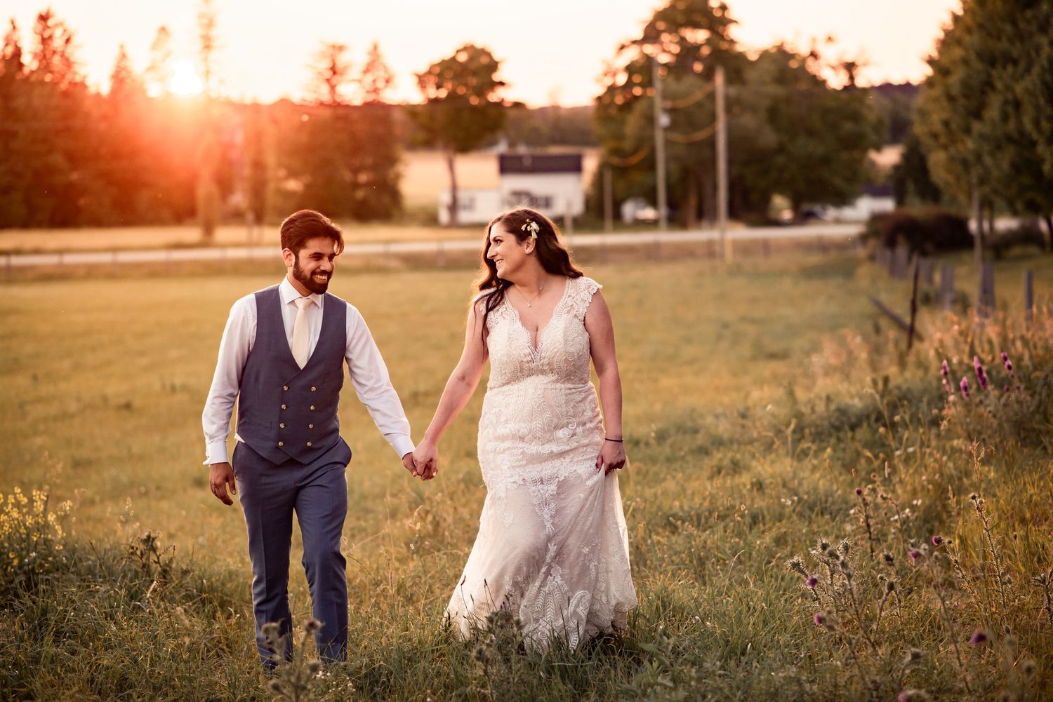 A couple walks hand in hand through a sunlit field at golden hour dressed in wedding attire.
