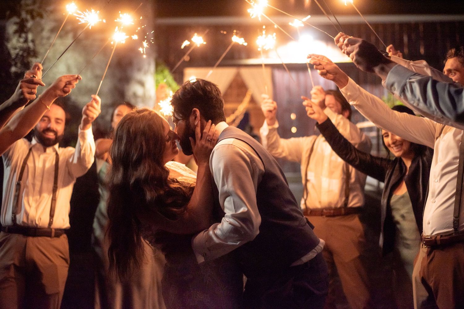 Guests dance and celebrate under warm string lights at an evening wedding reception with people raising their hands in joy.