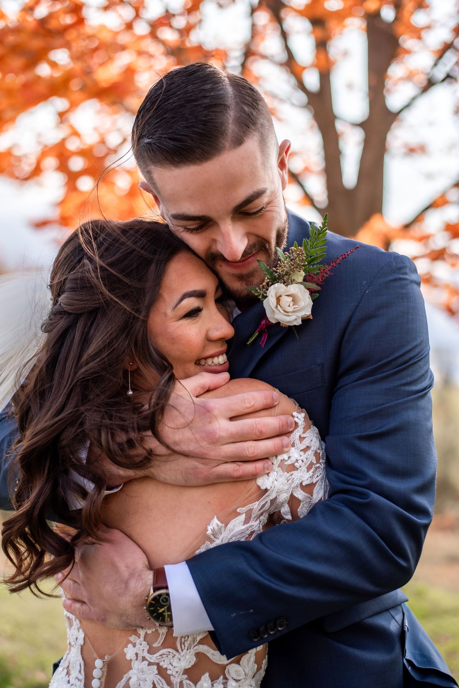 Happy couple embraces outdoors during autumn wedding with orange fall foliage in the background.