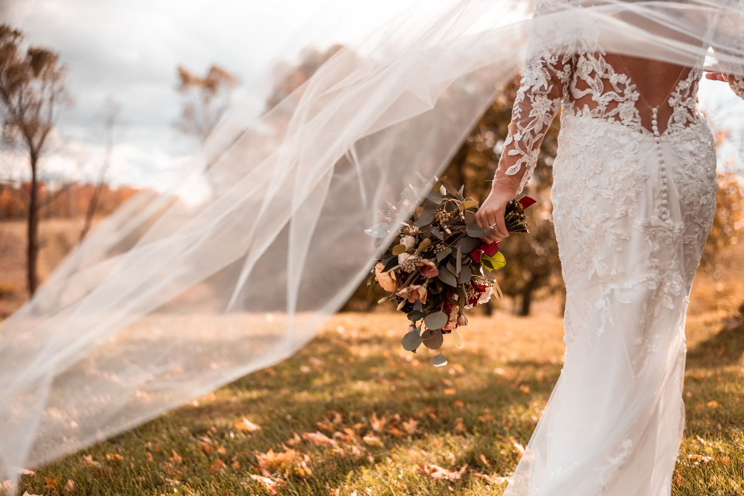 A flowing white wedding veil and lace dress detail with autumn flowers in a golden outdoor setting.