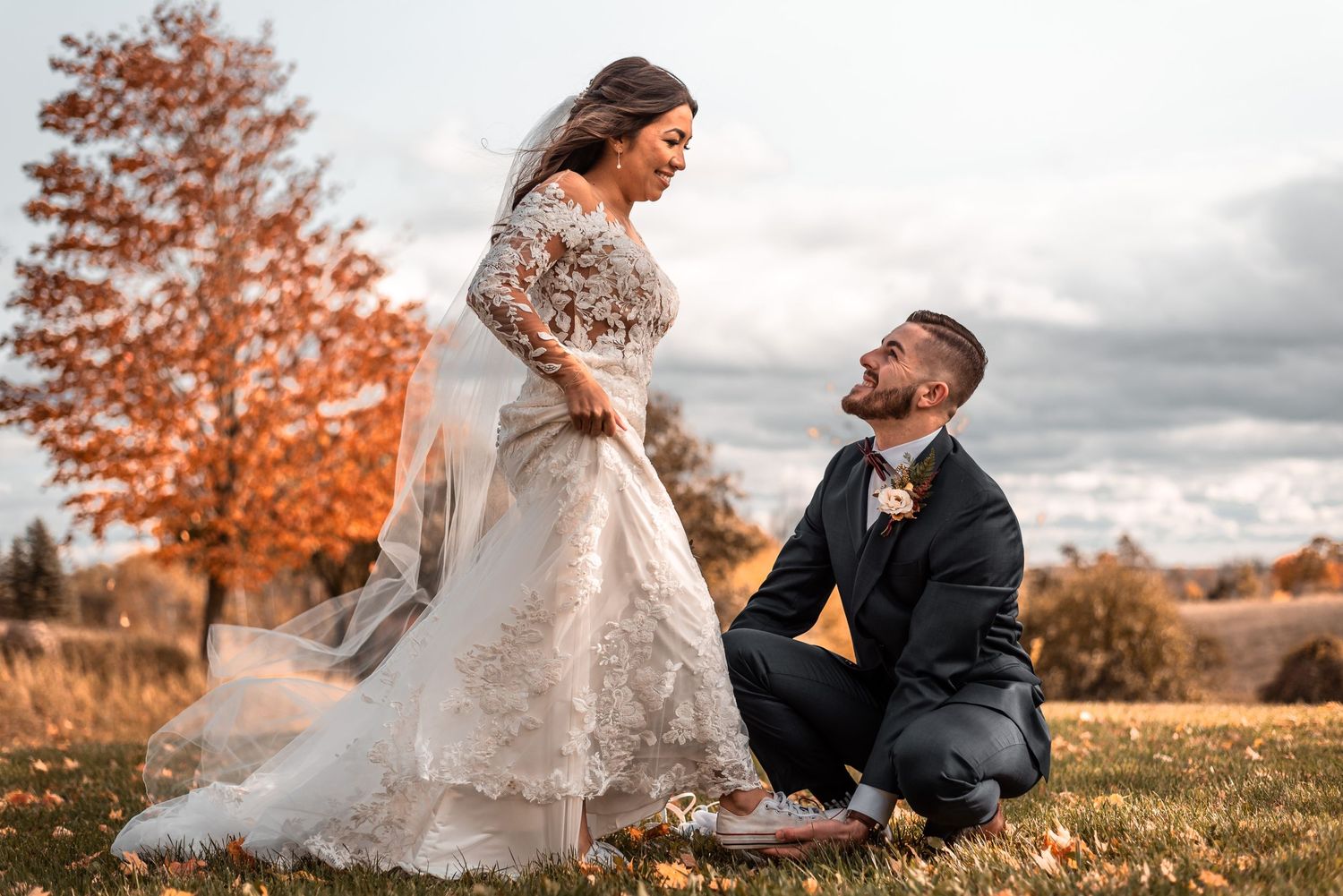 A romantic autumn wedding scene with couple in elegant attire kneeling on grass with orange fall foliage in background.