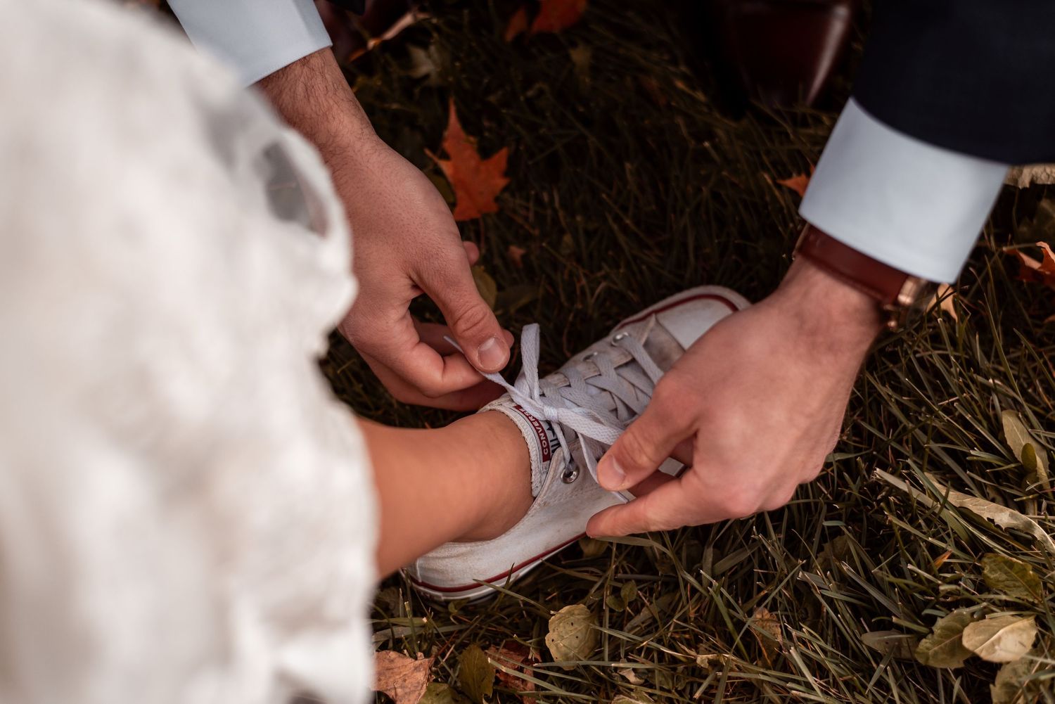 Close-up view of hands tying shoelaces on grey canvas sneakers against a ground covered in fallen leaves.