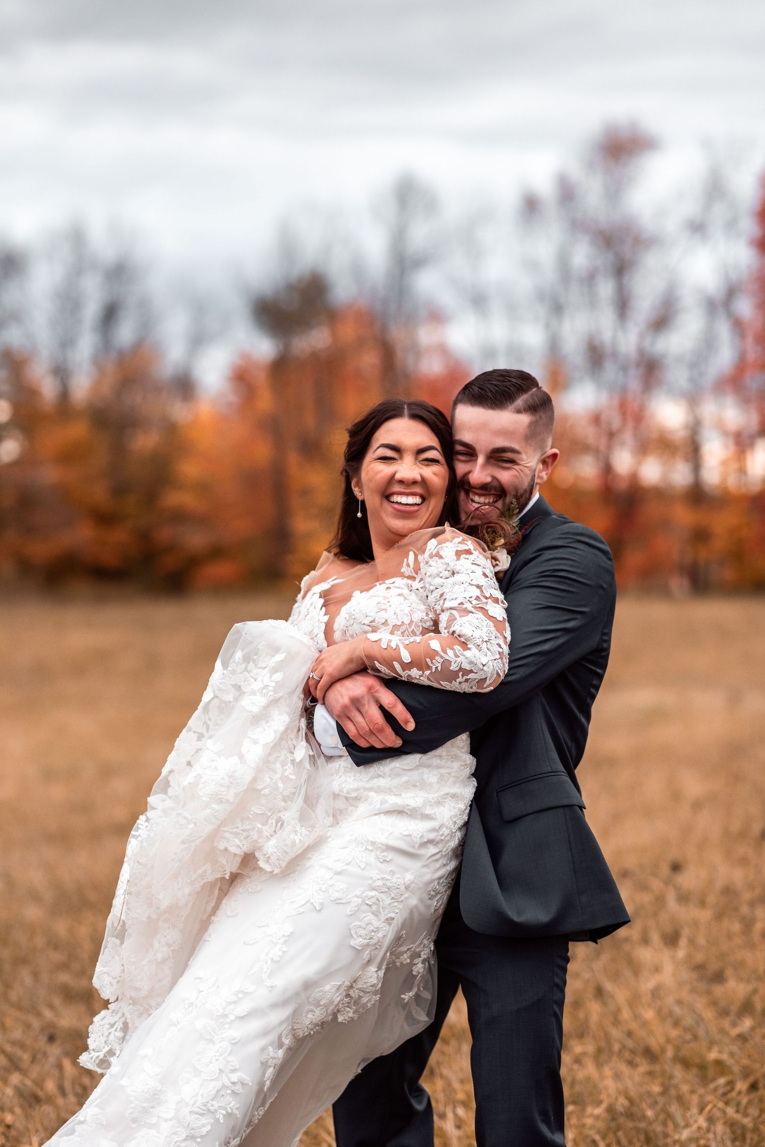 A joyful autumn wedding photo shows a couple embracing in a field with vibrant fall foliage in the background.