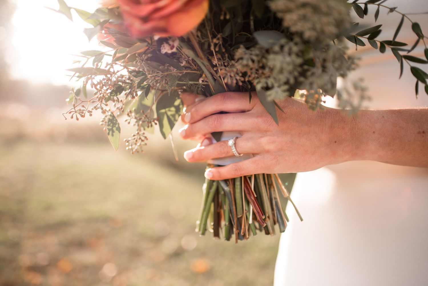 Close up of a bride's hand holding a bouquet of flowers with wedding ring visible during sunset.