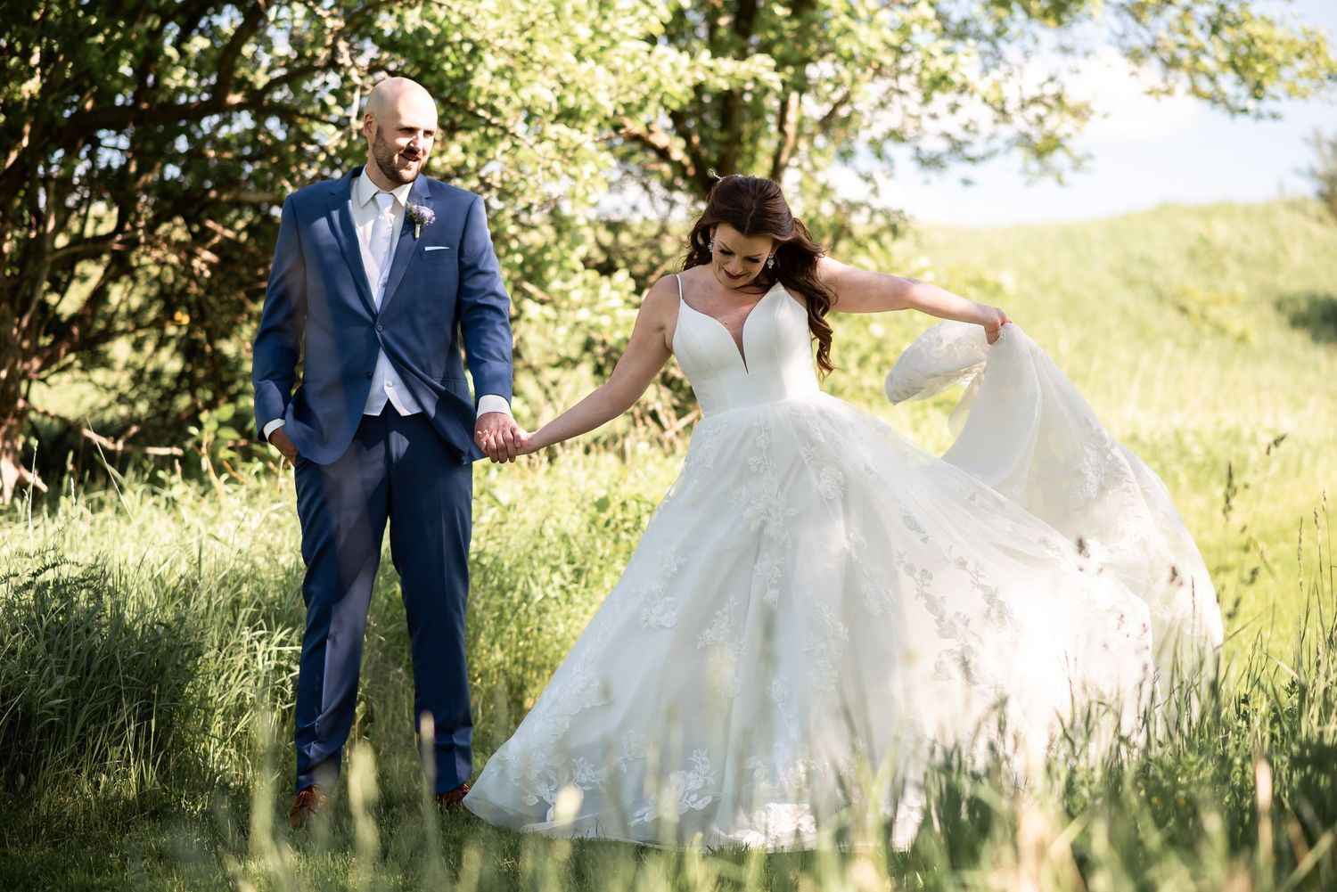 A couple in wedding attire share a playful moment in a sunlit meadow with long grass and trees.