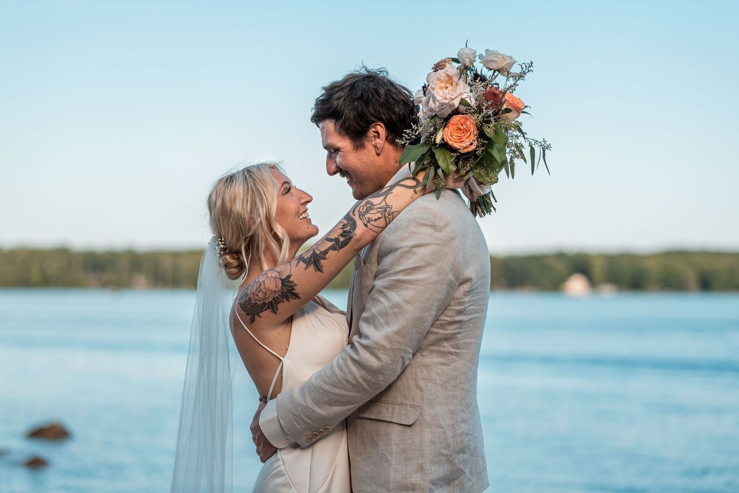 A romantic seaside wedding moment with a beautiful bouquet of peach and coral flowers against blue water.