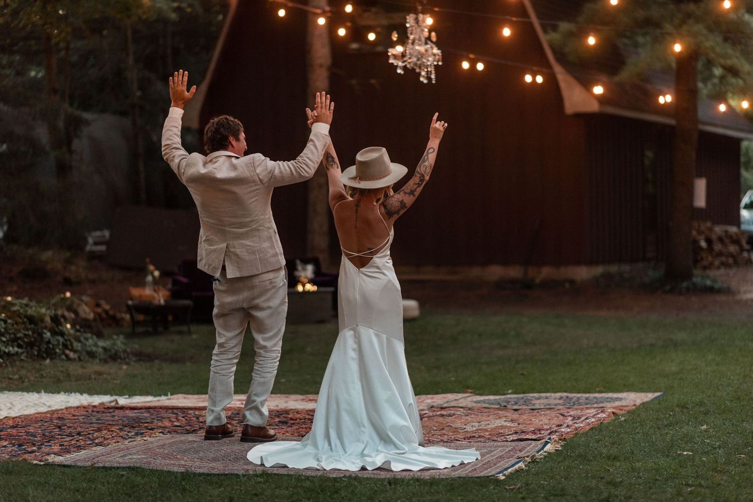 A couple celebrates their wedding with raised arms while standing on a rug at an outdoor evening reception with string lights.
