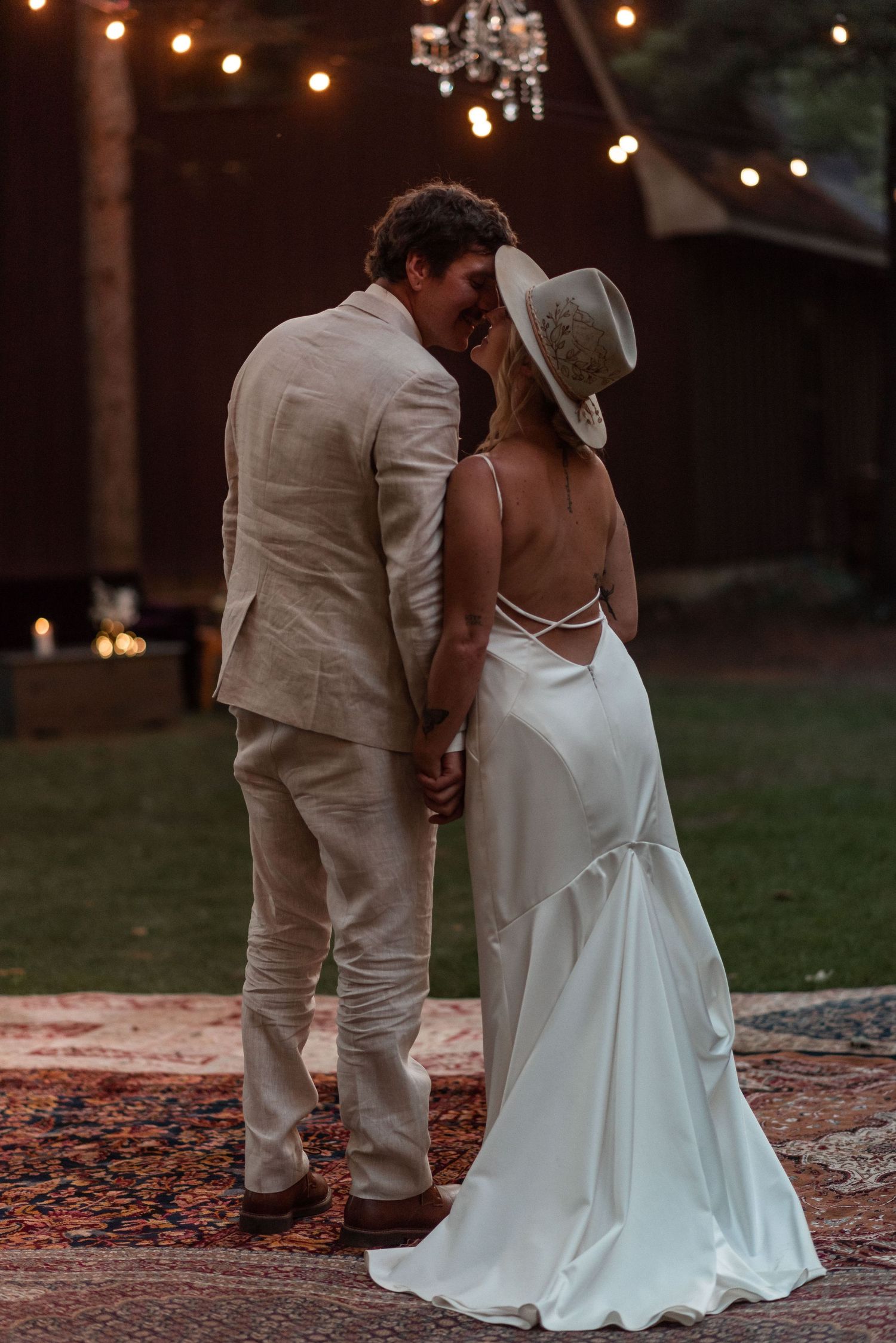 Newlywed couple shares intimate moment at dusk under string lights on a Persian rug outdoors.
