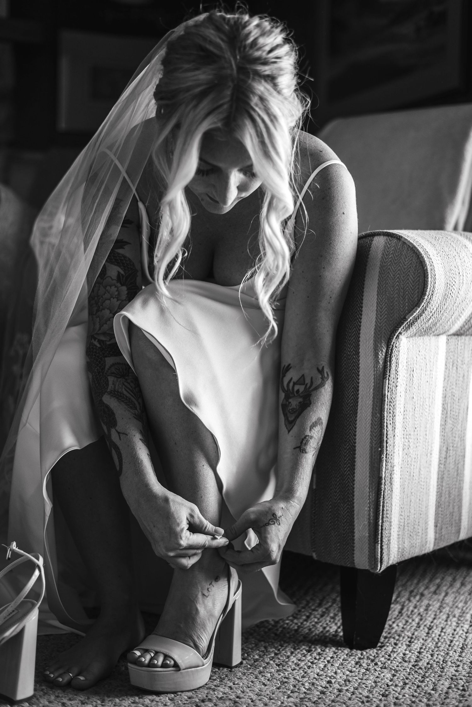 Black and white photo of bride in white dress and veil sitting on carpet adjusting her shoes before wedding ceremony.