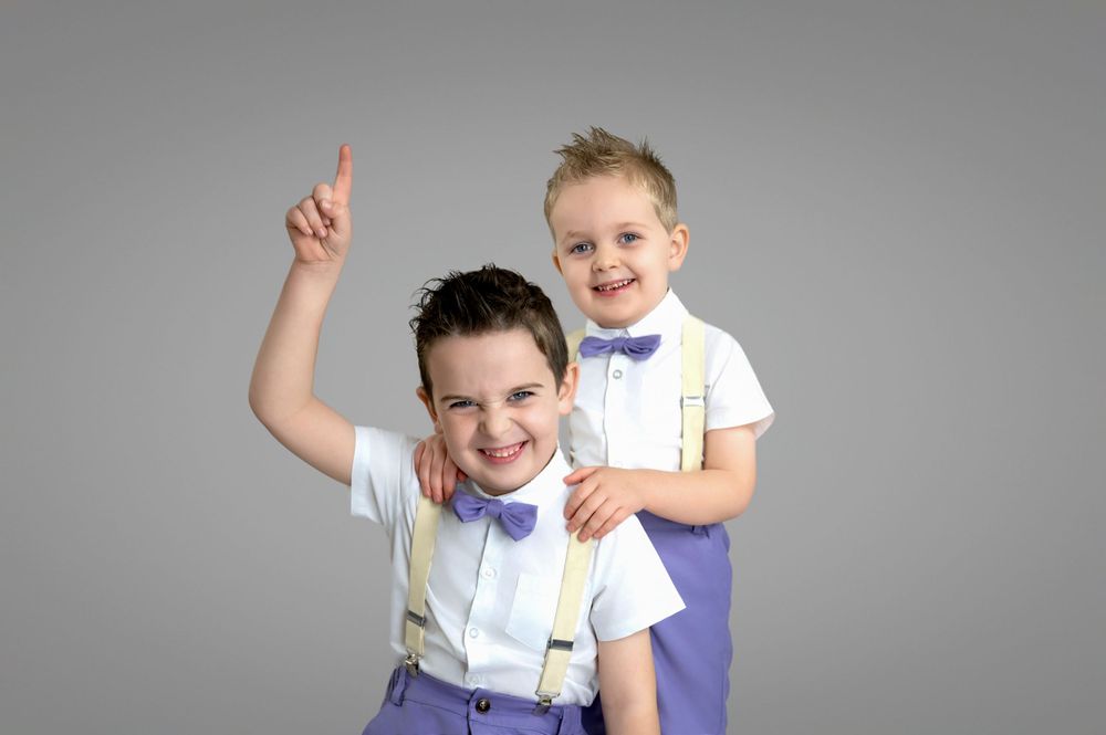 Two young boys in matching white shirts with suspenders and bowties pose playfully against a gray backdrop.