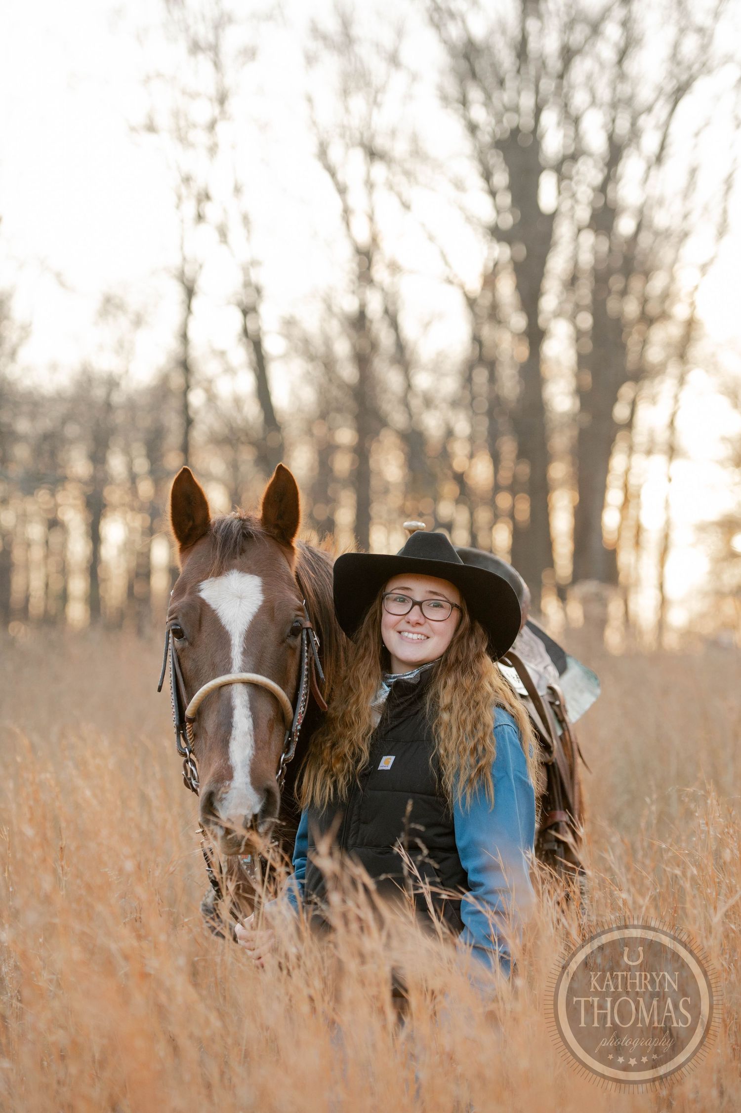 A rider in western attire poses with their horse among tall grass at sunset.