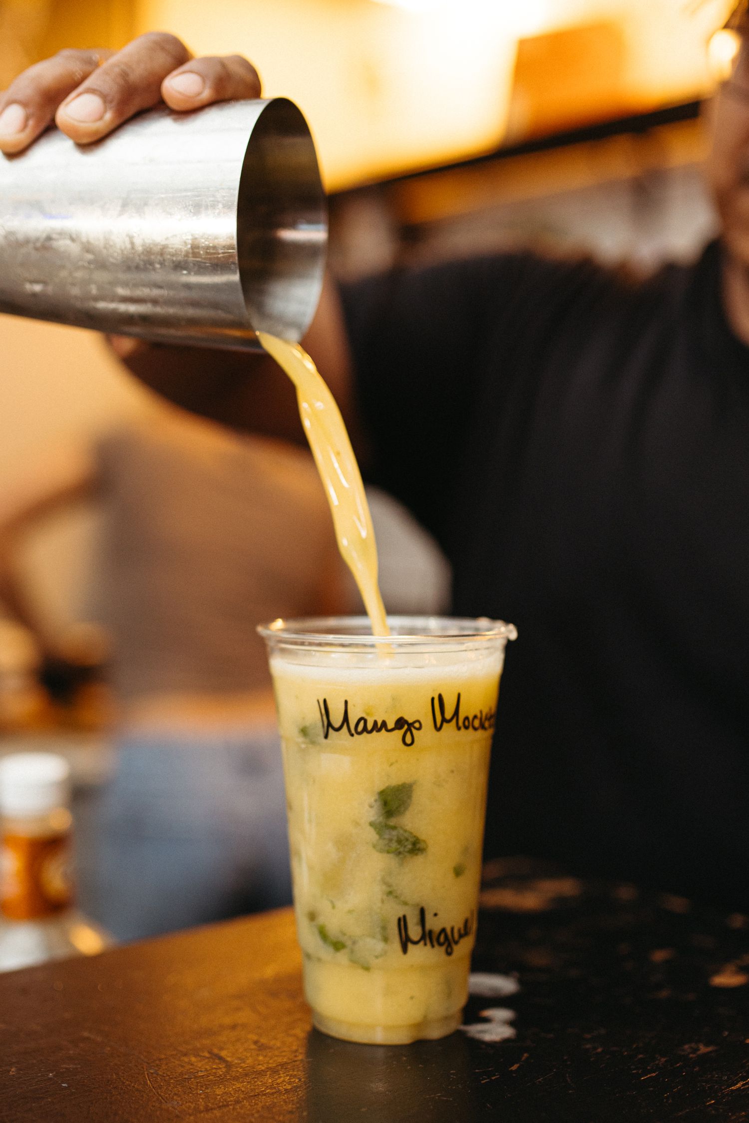 Yellow smoothie being poured into a clear plastic cup with green garnish on a dark wooden surface.