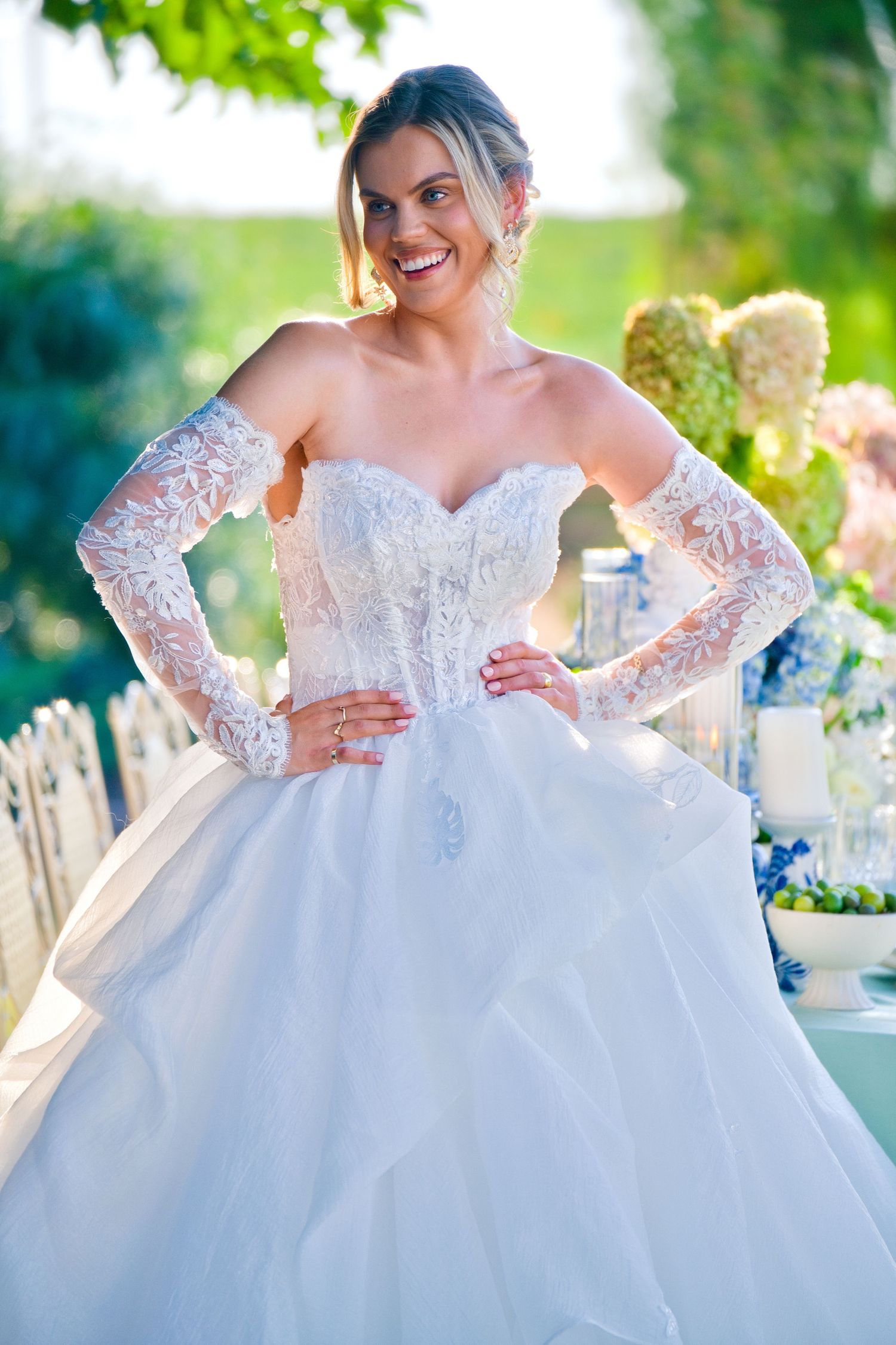 A bride poses in a stunning white ball gown with lace long sleeves and sweetheart neckline in a garden setting.