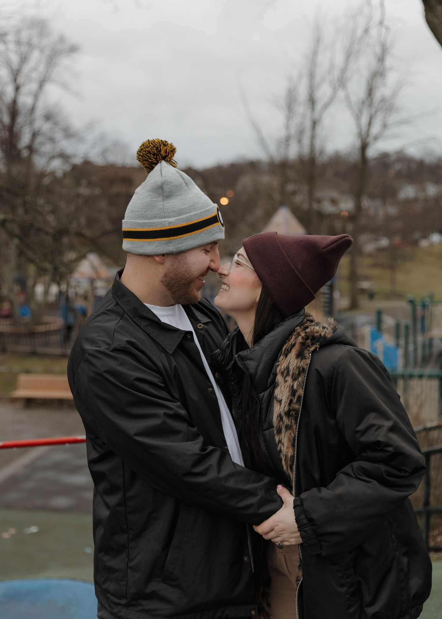 A couple wearing winter hats share an intimate moment at a park on an overcast day.