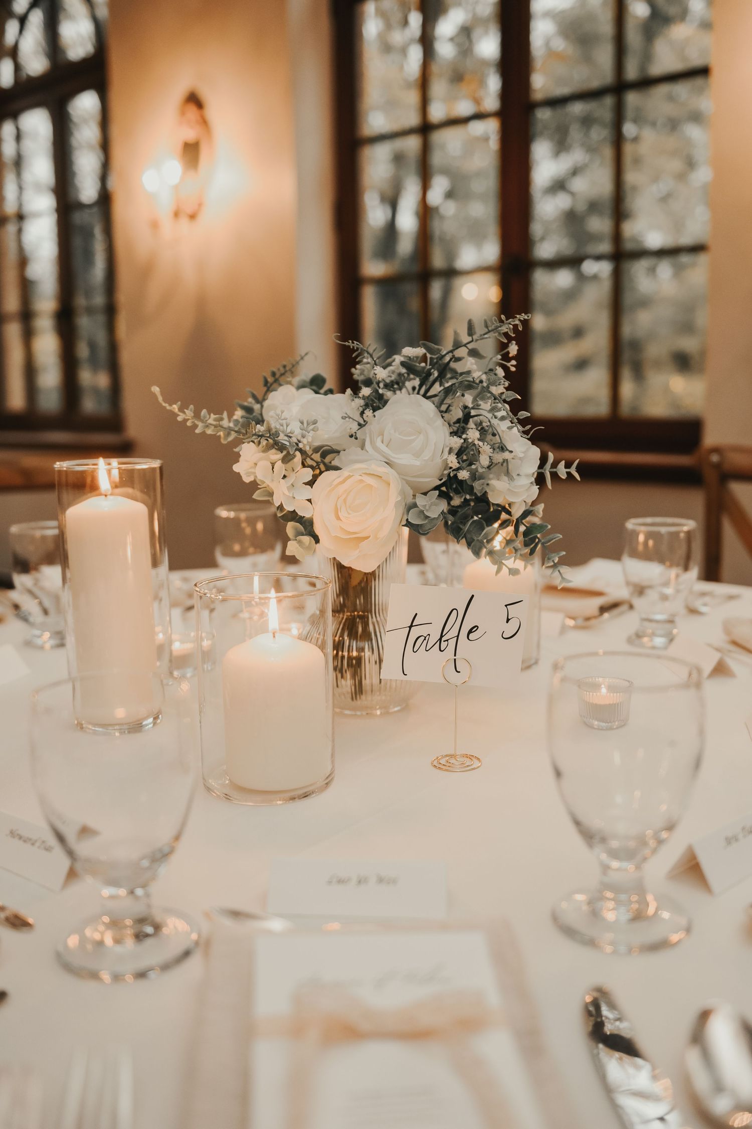 Elegant wedding reception table with white roses centerpiece, candles and crystal glasses in warm evening light.