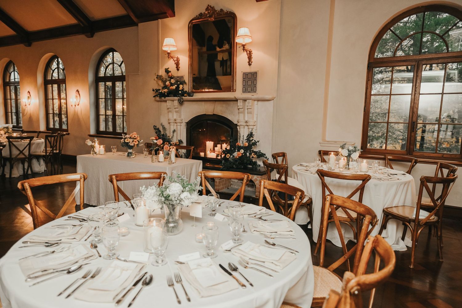 Elegant wedding reception setup with round tables, rustic wooden chairs, white linens and arched windows in a Mediterranean venue.