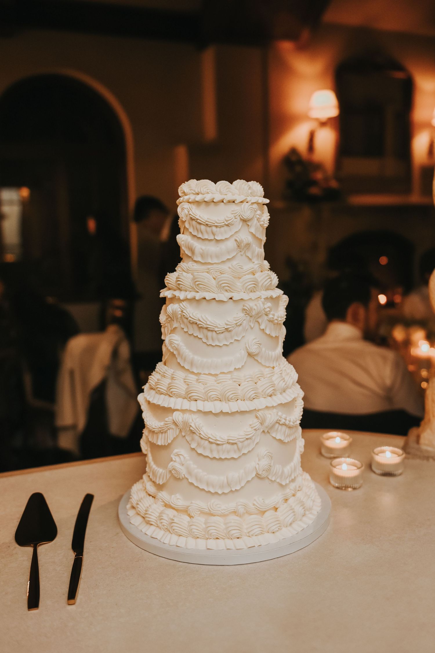 An elegant three-tiered white wedding cake with delicate piped swag details sits on a table with serving utensils nearby.