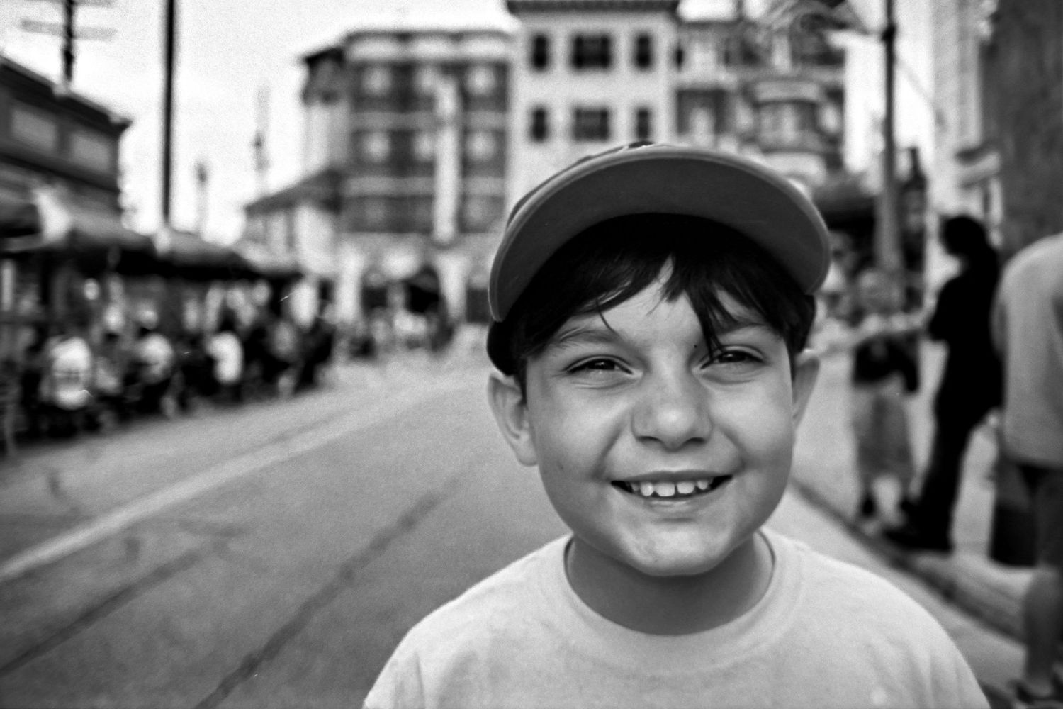 A young person in a baseball cap smiles brightly on a city street with buildings and pedestrians in the background.